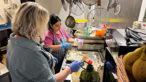 Two people wearing aprons and gloves prepare food together in a commercial kitchen, surrounded by hanging cooking utensils, ingredients, and stainless-steel work surfaces.