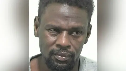 A police mugshot of Mohamed Harun, a man in his 30s with short black hair and a goatee. He is looking down and slightly to the right of the image.