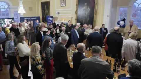 The Cromwell Museum A crowd of men and women in Huntingdon's town hall. They are looking towards Stuart Orme on the far right, who has grey hair and beard and is talking. There is a table with bottles of wine and glasses. There are portraits on the pale yellow walls. 