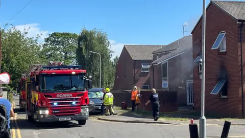The scene of a flat fire, two fire engines are on the left of the image in the middle of the road, fire service staff and a police officer can be seen on the street. To the left of the image is a two-storey block of fats, some of the brickwork is burnt around a ground floor window