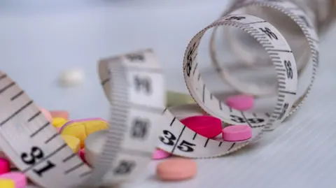 A white tape measure curled up with pink and yellow tablets on the surface.