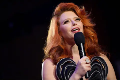 Getty Images Natasha Lyonne pictured with a microphone in a black dress