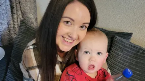 Alannah Alannah smiling with baby son Odin on her lap holding a bottle