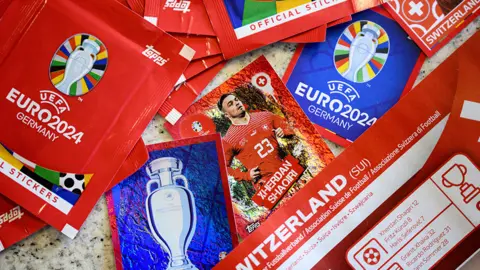 An artfully arranged pile of red sticker packets bearing the UEFA Euro 2024 Germany tournament name and trophy logo. In the centre, three stickers are visible - two show the trophy logo and tournament name on a blue background, the third shows Switzerland player Xerdan Shaqiri.