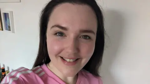 Emma Bowell Emma Bowell smiling for a selfie, with a white wall behind her. She is a white woman with long dark hair and is wearing a pink top with three white stripes on the shoulders.