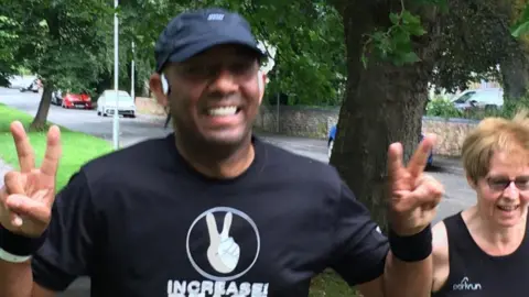 BBC A man wearing a black t-shirt and cap doing a peace sign with his hands