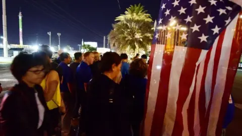 AFP People look on near a US national flag at a makeshift memorial near the Mandalay Hotel on the Las Vegas Strip (03 October 2017)