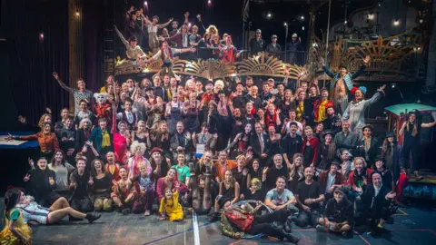 Andre Pattenden A massive group of performers in circus attire of all ages smiling with hands outstretched at the camera