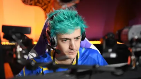 Getty Images Ninja, the well-known streamer, looks like he's concentrating on a match as the orange glow of a gaming room emanates from behind him. He's wearing a large gaming headset with padded earcups and what looks like an American hockey or football style shirt in blue with yellow lines criss-crossing at points. His trademark spiky hair is coloured a teal/blue shade on this occasion.