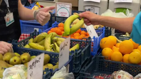 BBC a hand picks two bananas from a table with boxes of bananas, pears and oranges on it