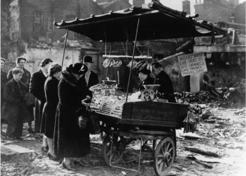 Getty Images People queuing to buy oranges and bananas at a stall in bomb-wrecked London, during the Blitz, 23rd November 1940. A sign on the stall reads: 'Hitler's bombs can't beat us. Our oranges came through Musso?s lake'. Musso?s lake was service slang for the Mediterranean sea. (Photo by Keystone/Getty Images)