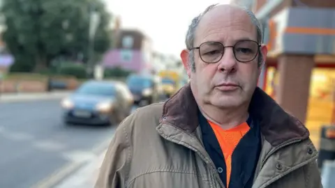 Stuart Woodward/BBC A white man looking at the camera. He is wearing glasses with a black frame a coat and an orange shirt.