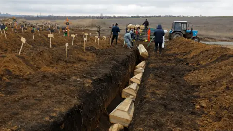 Reuters Coffins are placed into a long trench outside Mariupol in February 2023