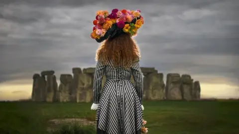 Andre Pattenden The back of a woman with long hair wearing a bright headdress with lots of pink and orange dahlias standing in front of Stonehenge with a grey sky.