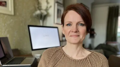 BBC Anna Rowe in a beige jumper sitting by a desk