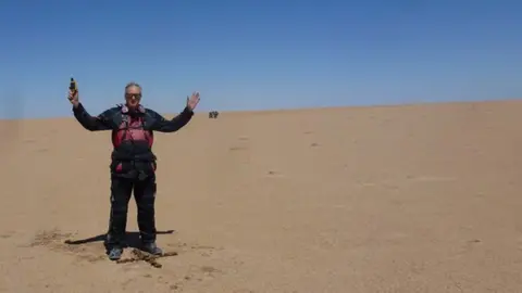 John Abell A man stands in the middle of a barren desert, above an 'X' carved into the sand