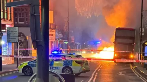 X/RodCam24 View of a street in Wimbledon filled with smoke and the glow of flames behind a bus