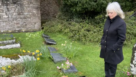 BBC Jacki Sutton looking down at graves in a cemetery wearing all black