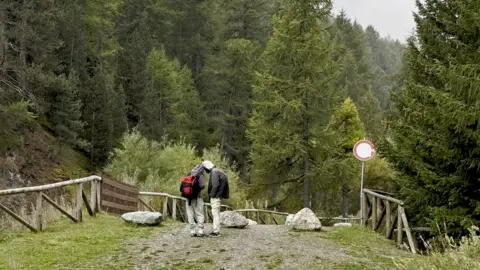 Two young men stand on a mountain gravel track on the Italian side of the French-Italian border, wearing trainers and carrying a small bag