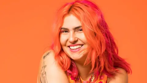 Ruby Rare Ruby Rare, a young woman with dyed orange and pink hair, smiles at the camera. Ruby wears pink eyeshadow and lipstick and wears her hair loose around her shoulders. Her arms are folded across her front, revealing a fine-line tattoo on her right shoulder and down her arm. She wears a sleeveless orange and pink top and is photographed in front of an orange background,