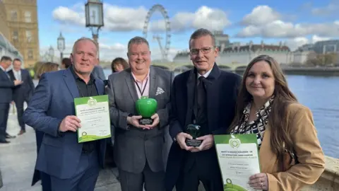 Waste to Wonder Four people standing with a view of London behind them, including the London Eye, holding green apple ornaments and certificates