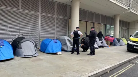 Mariam Issimdar/BBC Image showing a female officer bent down talking to someone inside a tent on the street