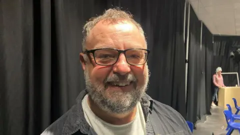 Tom Percival/BBC Man with grey hair and beard wearing glasses in a rehearsal studio