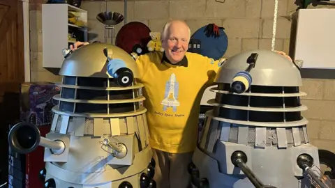 Dave Latham wearing a bright yellow sweater standing between the two recovered daleks