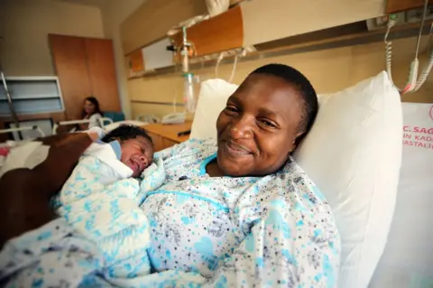 Getty Images Cameroonian mother Agol Tchabda, who gave birth on irregular migrants' boat, lies on a stretcher as she holds her baby at a hospital after the migrants were rescued by the Turkish Coast Guard units in Aydin, Turkiye on Junly 01, 2022.