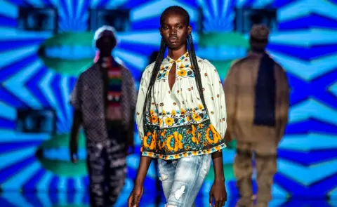 EPA Majda John Peter on the runway. There are two models behind her. She is wearing jeans, a printed top and braids.