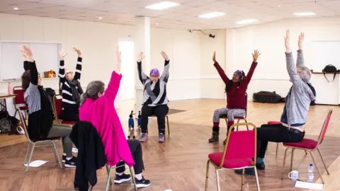 Leyla Ozkan People dancing in their chairs, arranged in a circle, in a hall