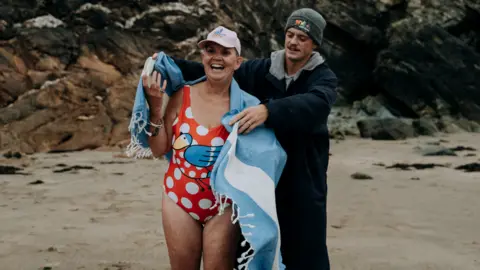 Ella Richardson A man wrapping a towel around an older woman in a swimsuit on a beach