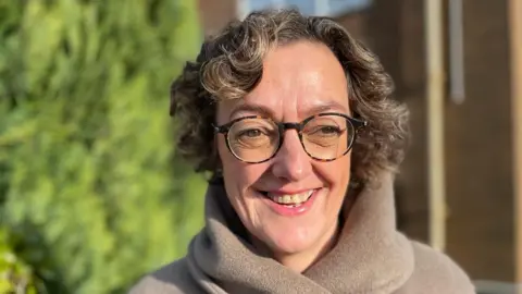 Dr Williams is wearing a brown woollen coat and dark framed glasses. She has short, brown curly hair and is smiling. She is stood outside Velindre Cancer Care centre.