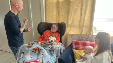 BBC A man speaking to a girl watched by a woman holding a baby