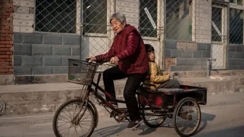 Getty Images An elderly woman drives a tricycle as a boy sits on a back seat on a street in Beijing.