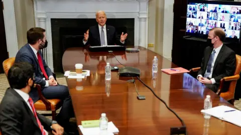 Getty Images President Biden and three advisers sit around a table as business leaders join by video link