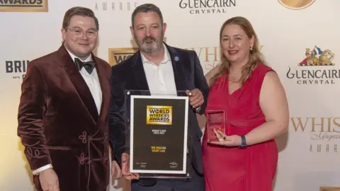 Two men and one women. at an award ceremony. The man in the middle is wearing a shirt and a blazer while holding the award/ On his left is another man smiling wearing a bowtie and on his right a woman is smiling while wearing a pink dress