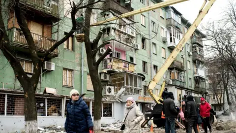 Getty Images People walk on the street as emergency teams check a residential building damaged by a Russian missile attack in the city of Zaporizhzhia, southern Ukraine. Photo: January 2023