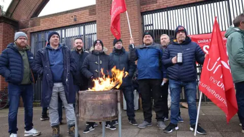 PAcemaker men keeping warm around a fire while flying Unite the Union flags