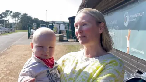 BBC Heidi Baker, who has blonde hair, is smiling while looking off to the side. She is wearing a light-coloured sweater with a yellow-green pattern. She is holding a baby, dressed in a white outfit with a red bandana bib.