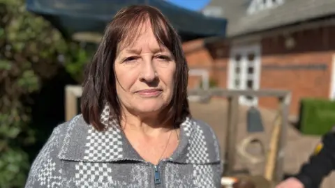 A woman with short brown hair and a fringe is looking at the camera with a slight smile on her face. She has a nose piercing and is wearing a silver necklace. She is wearing a grey zip up jumper with a collar. The sun is shining and there is a building with a window in the background.
