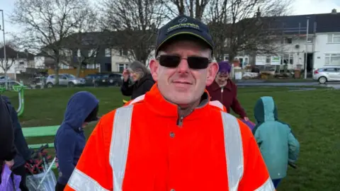 Wearing a high vis jacket, a dark baseball cap and rectangular sun glasses, Martin Hoe looks at the camera. He stands in a green space with two children and two adults visible behind him. The young children both have hoods up, with the sky full of grey clouds.