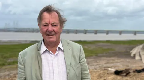 Dr Andrew Garrard wearing a sage blazer and pink patterned shirt stood on the banks of the River Severn in front of the first Severn crossing. 