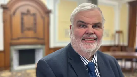 Dave Thomas is wearing a blue and white striped shirt and a navy blue tie and jacket and is standing in a committee room
