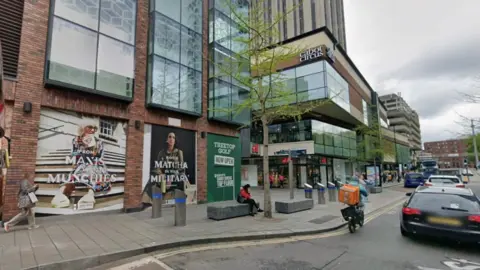 A Google Street View image shows the scene on Penn Street through Cabot Circus. On the pavement a person sits on a public bench in front of a shop window covered with an advert. High up on a building behind them to their left, a sign reads "Cabot Circus". 