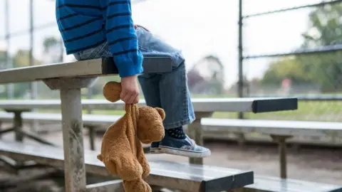 File photo of a child dressed in a blue T-shirt and jeans, whose face is out of shot. The child is carrying a brown teddy bear and sites in the stand at a sporting venue,