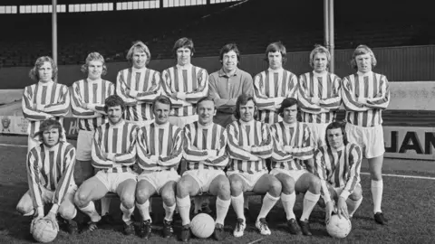 Getty Images A black and white photograph of an old Stoke City FC team pictured on the pitch in a stadium. There is a line of players standing at the back with their arms folded and a line at the front either sitting on chairs or kneeling on the ground.