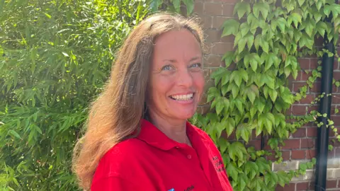 Clare Worden/BBC Vicki Bailey is standing in front of a wall covered in green leaves. She has shoulder-length brown hair and is wearing a red polo shirt. 