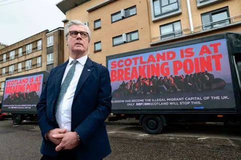 Getty Images Reform’s Scottish leader Malcolm Offord wearing a blue suit, white shirt and green tie, attends a party election event where he stands in front of a van with the message 'Scotland is at breaking point' written on the side.
