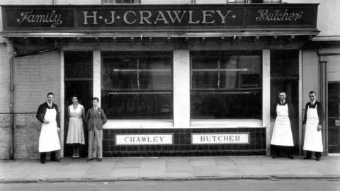 Other A black and white photo of a butchers in the 1920s, with five people outside, four men and a woman, split into a group of three at one end and two the other, three of the men wearing white aprons. Between them, on the wall of the building is a sign saying "Crawley Butcher".